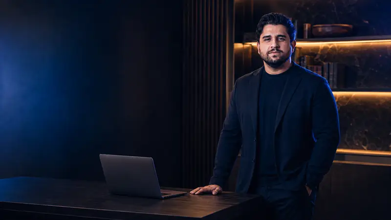Rodrigo Neves em pé ao lado da mesa do escritório, com estante ao fundo